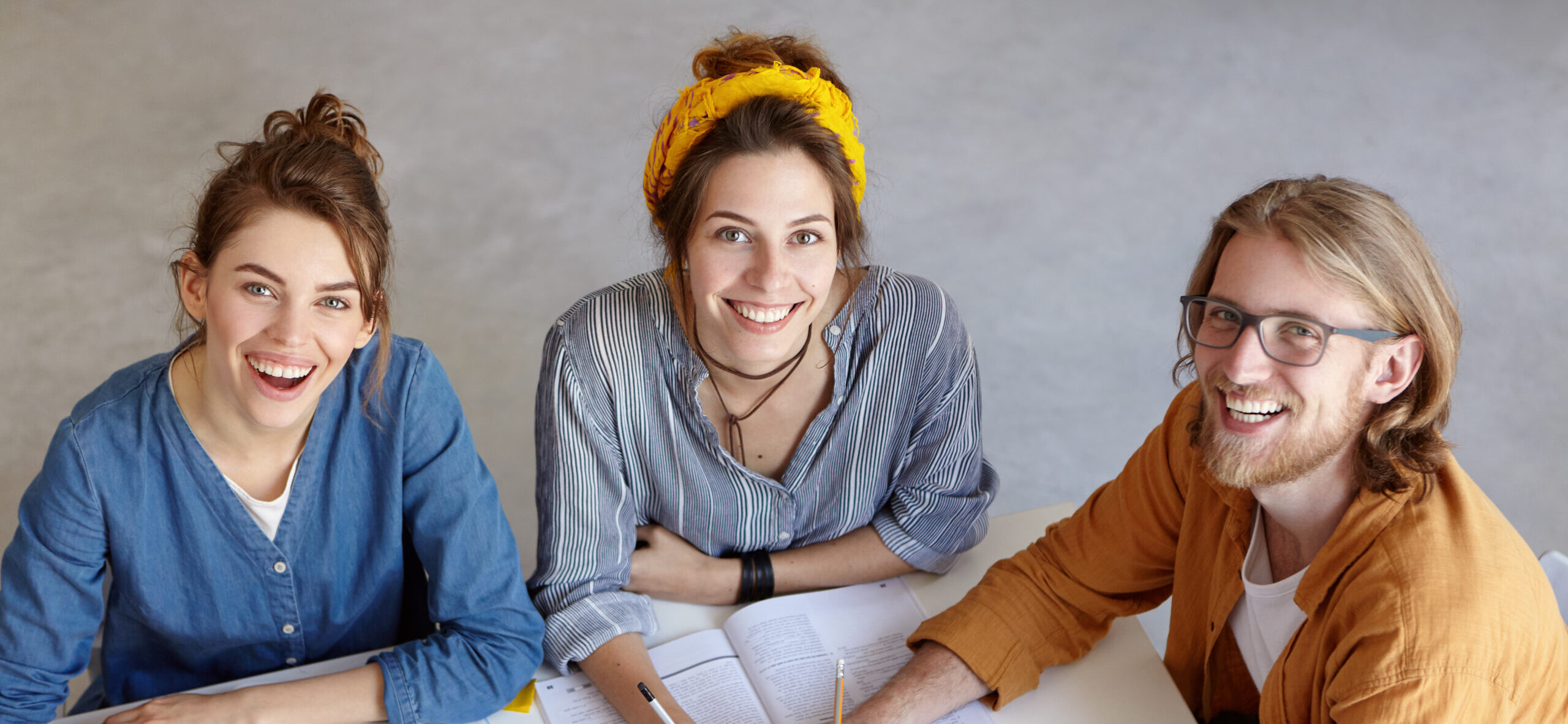 Positive co-workers smiling at camera with delightful look sitting at their work place. Three researchers reading new publications. Creative students being happy to study together. Education concept