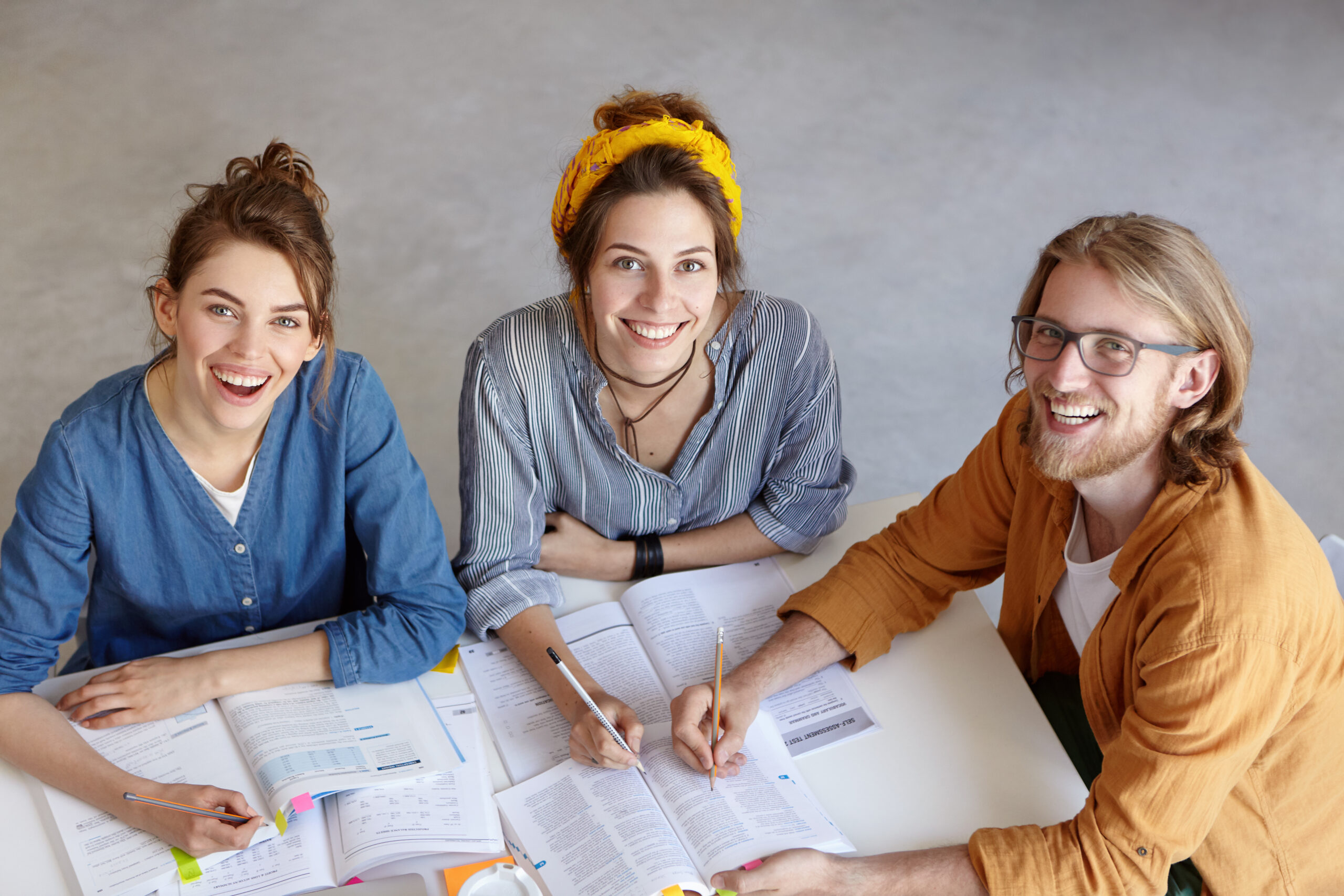 Positive co-workers smiling at camera with delightful look sitting at their work place. Three researchers reading new publications. Creative students being happy to study together. Education concept